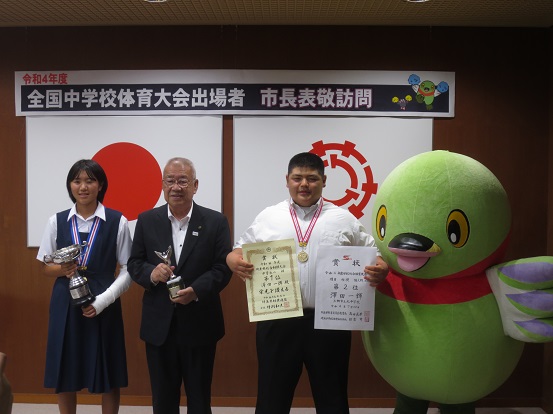 壁に「令和4年度 全国中学校体育大会出場者 市長表敬訪問」のボードが設置されている部屋で、メダルを肩にかけて表彰状やトロフィーを持った生徒2名と一緒に市長とかいちゃんが並んで記念撮影をしている写真
