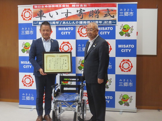 三郷市のバックボードに「車いす寄贈式」の横断幕が設置されている部屋で、車いすを真ん中に置いて記念撮影をする菅川越谷支社長(左)と木津市長(右)の写真