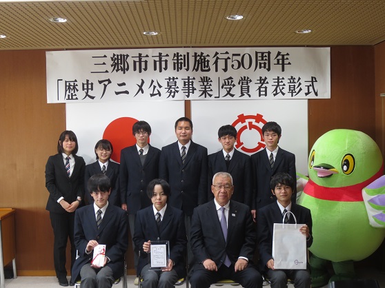 三郷市市施行50周年記念「歴史アニメ公募事業」受賞者表彰式と書かれた横断幕と国旗、市章の前で、目録や盾を持つ9名の県立三郷工業技術高等学校 情報電子科3年映像班の皆さんとかいちゃん、と木津市長の集合写真