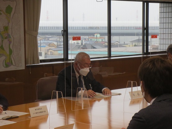 窓が広がりパーテンションで区切られた会議室で、自席に座り挨拶をしている三郷市長と席に座って聞いている関係者の方々の写真