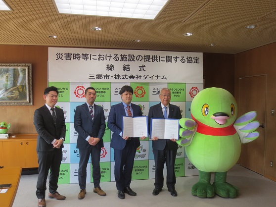 「災害時における施設の提供に関する協定締結式」と書かれた三郷市のバックボードの前で、3人の株式会社ダイナムの男性と三郷市長、かいちゃんが横1列に並び締結書を持って一緒に記念撮影をしている写真