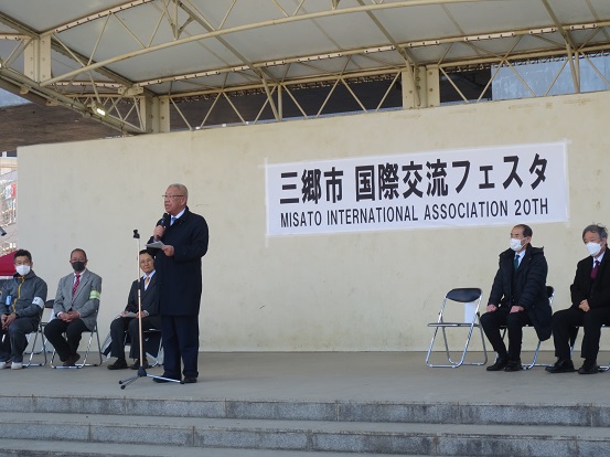 におどり公園で「三郷市国際交流フェスタ」と書かれたステージでスタンドマイクの前に立ち挨拶している三郷市長と、パイプ椅子に座っている6人の関係者の方々の写真