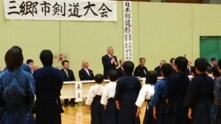 三郷市剣道大会と書かれた横断幕の下に設置された来賓席中央の席から立ちあがってマイクを持ち挨拶を行っている市長の写真