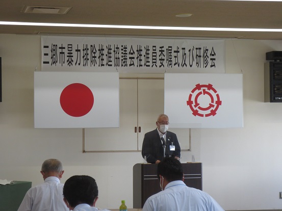 「三郷市暴力排除推進協議会推進員委嘱式及び研修会」の横断幕が貼られている部屋で、市長が演壇に立って話をしている写真