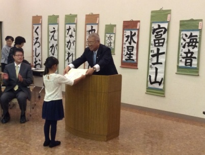8枚の書道の作品が掛けられた壁の前に設置された演壇を挟んで市長が女子児童に賞状を手渡している表彰式の様子の写真