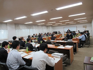 地区協議会で、前に立って話す男性と座って聞く大勢の参加者達写した会場内の様子の写真