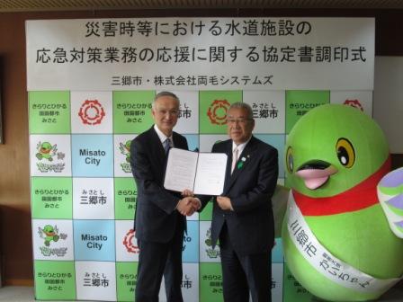 「災害等における水道施設の応急対策業務の応援に関する協定書調印式」とかかれたパネルの前で市長と両毛システムズの代表の方が一緒に調停書を持ち、三郷市マスコットキャラクターかいちゃんと一緒に記念撮影している写真