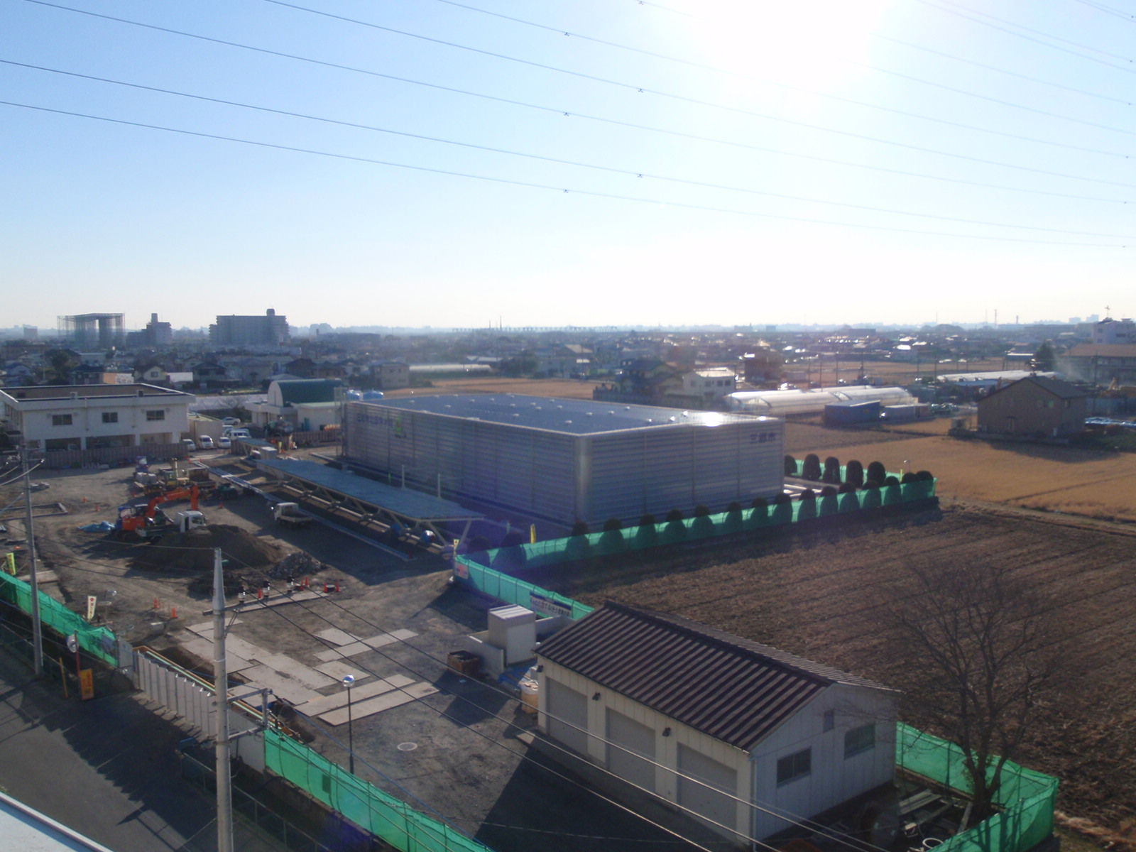 敷地の右半分に完成された四角状の建物が建ち、左半分のスペースでは盛られた土でショベルカーが作業をしている写真