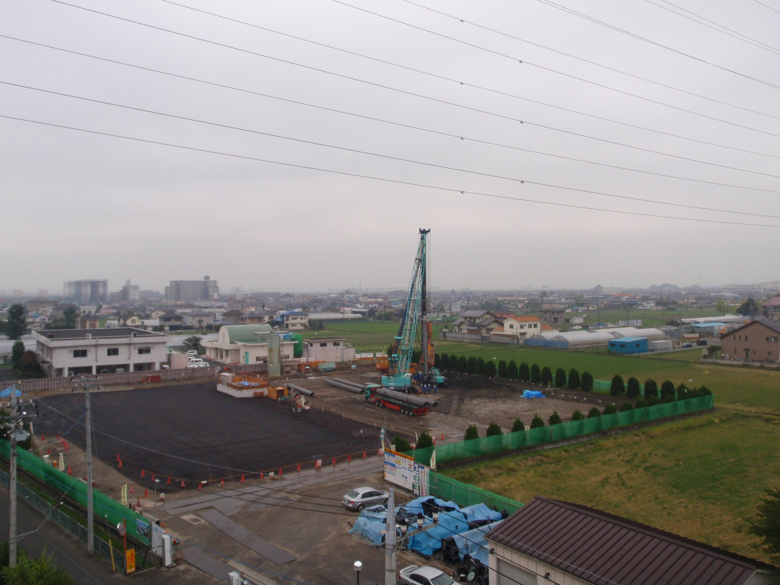 工事用フェンスで囲まれた敷地の奥に緑色のクレーン車やトラックが作業をしている写真