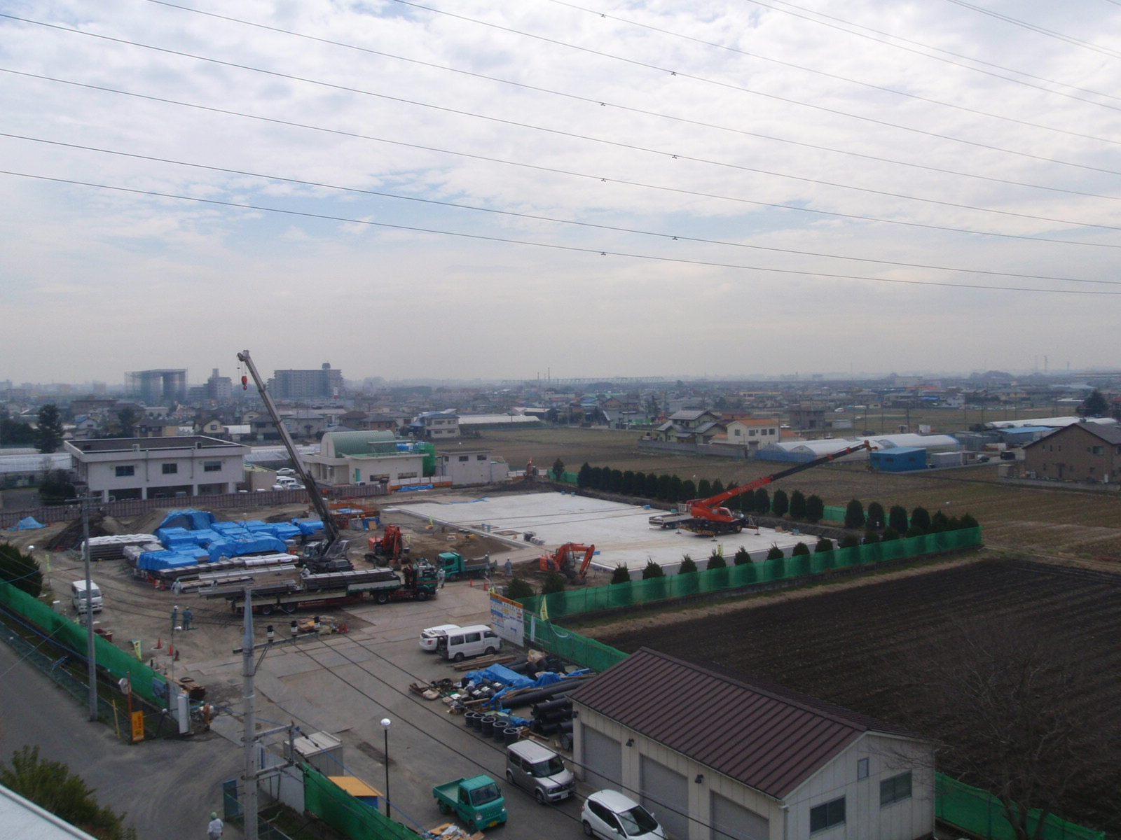 多くの工事用材料が置かれた敷地で2台のクレーン車や多くの重機が作業をしている写真