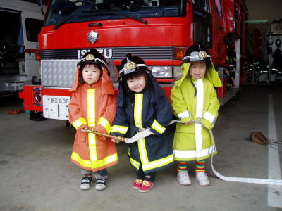 3人の子どもが子ども用の消防服を着て、消防車の前で小さなホースを持っている写真