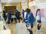投票箱に投票用紙を入れている1名の女子児童と、後方で記載や説明を受けている女子児童たちのデザート選挙の様子の写真