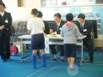 長机と椅子が設置された会場で3名の職員の方が座り、受付をしている2人の男子児童の写真