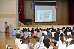 学校の体育館で多くの保護者に栄養教諭がスライドを見せながら、食育の講話を行っている写真。