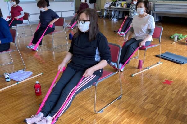数名の女性たちが椅子に座り両手で紐を持ち、足にひっかけて伸ばしている写真