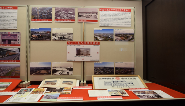 台の上の赤いマットに三郷市庁舎パンフレットや三郷市勢要覧などが置かれ、正面ボードに市庁舎の移り変わりの写真や空から見た市役所周辺の写真が展示されている写真