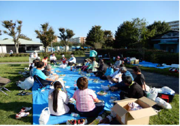 早稲田公園テニスコート横の芝生にブルーシートを敷き20名程が輪になって団らんしている懇親会の様子の写真