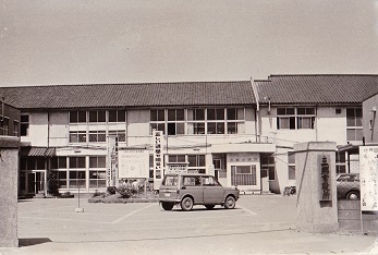 昭和50年(1975年)ごろに撮影された、二階建ての建物の前に1台の車が駐車し、その手前の門に「三郷市役所」の表札が掲げてある旧市役所庁舎のモノクロ写真