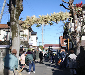 丸太と藁を使って作られた蛇が、道路の両脇に生えた2本の木に掛けられており、木の周辺を住民が掃除している写真