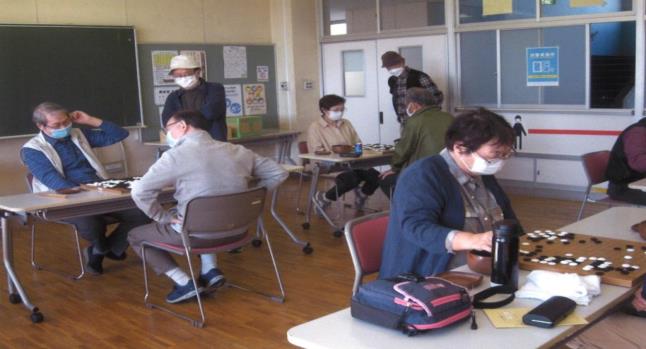 奥で2組が対局を行い、それぞれの机の横に男性が立ち対局を見ている様子の写真