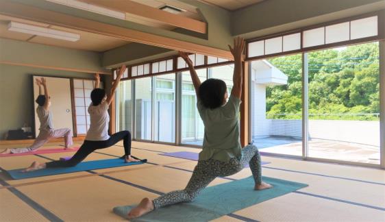 ヨガマットの上で、窓側を向き、右足は前、左足は後ろで両手を上に上げてポーズを取っている3名の女性の写真