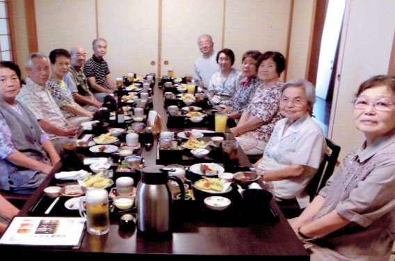 奥に長く並べられた長机の上に黒いお膳にのった料理と飲み物の入ったグラスが並び、机の両側に6人ずつ椅子に座った高齢の男女がカメラの方を見ている懇親会の様子の写真