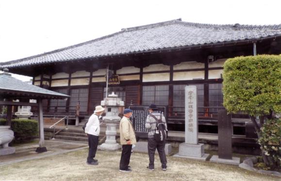 3人の男性が瓦屋根で横長の迎攝院(本堂)を見学している三郷市内史跡巡りの写真