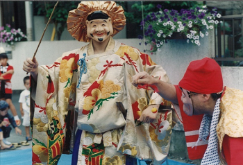 恵比寿様のお面をかぶった人が釣り竿を持ち、釣り糸を持つ右側の男性とともに恵比寿の鯛釣りを演じる写真