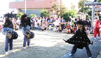 大勢の観客が見ているなか頭に獅子頭をつけた2名の踊り手が腰につけた太鼓を打ち1名が両手と両脚を広げて舞っている様子の写真