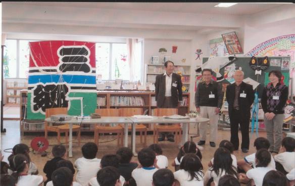 手前に小学生が並んで座り、奥に三郷と書かれた大きな凧と4人の会員が並んでいる写真