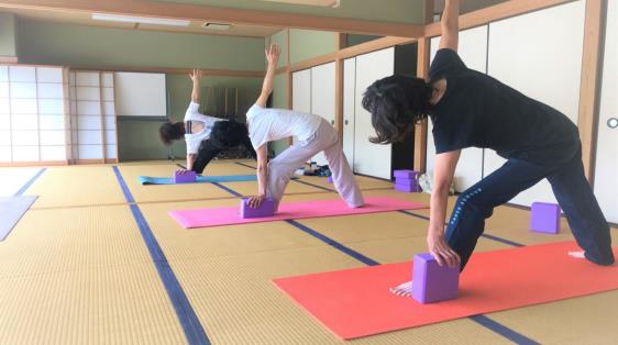 左足を前に出し、左手はブロックの上に、右手は真っすぐ上に上げて三角ポーズを取っている3名の女性の写真