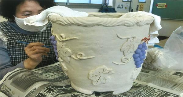女性が植木鉢の形に成形され葉っぱやぶどうがデザインされた作品に、色を塗っている写真