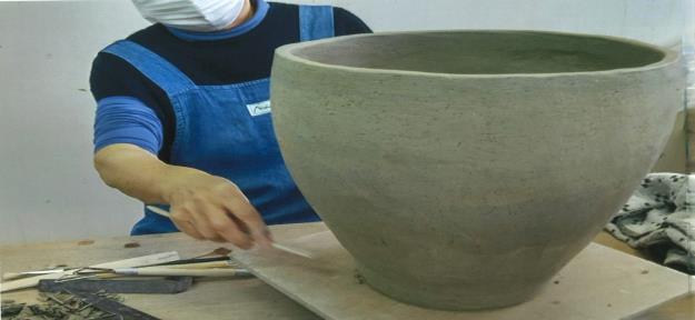 青いエプロン姿の女性の前に大きな粘土の器が置かれ、ヘラのような道具を使って作業をしている様子の写真