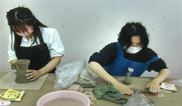 青いエプロンをした女性は粘土を棒に丸め、黒いエプロンの女性は器を作り陶芸を楽しんでいる写真