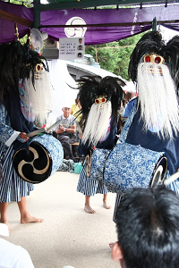 舞台の上で白と青色のストライプ柄の着物を着用し頭に獅子頭をつけた3名の踊り手が腰につけた太鼓を打ち舞っている様子の写真