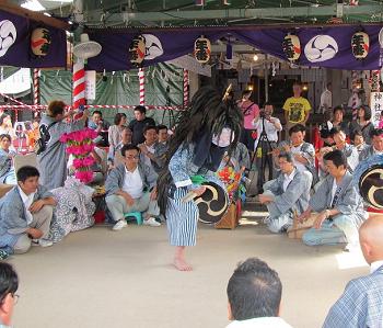 神社の拝殿前の舞台で周りの笛と太鼓の演奏に合わせて白と青色のストライプ柄の着物を着た踊り手が頭に獅子頭をつけ腰につけた太鼓を打ち舞っている様子の写真