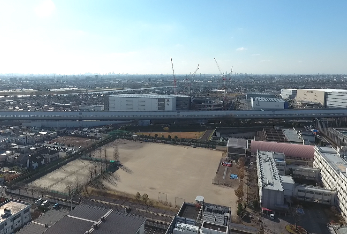 令和3年(2021年)に撮影された、右手に三郷高校、左手に広い運動場がある三郷高校とその奥でいくつものクレーン車が工事をしている写真