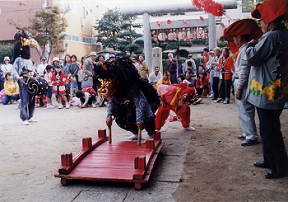 神社の境内に設置された小さな朱色の橋の上を頭に角が生えた獅子が両手に握った棒を橋について渡っている舞いの様子の写真
