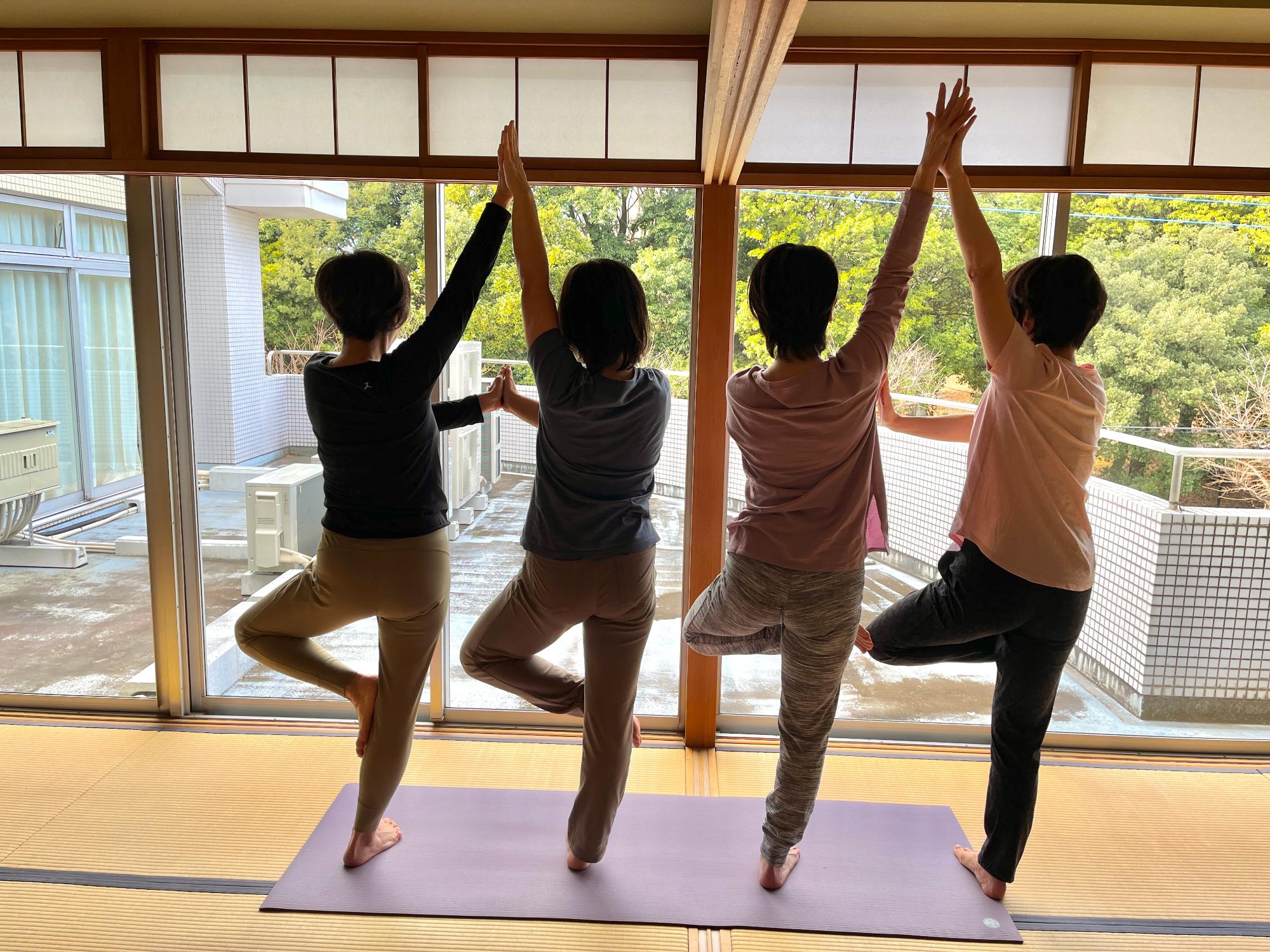 ヨガマットの上で、両手を合わせて上にあげ、片足で立ち木のポーズをしている3名の女性の写真