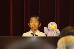 コバトンのマスコット人形がある演台のマイクで発表をしている制服を着た女の子の写真