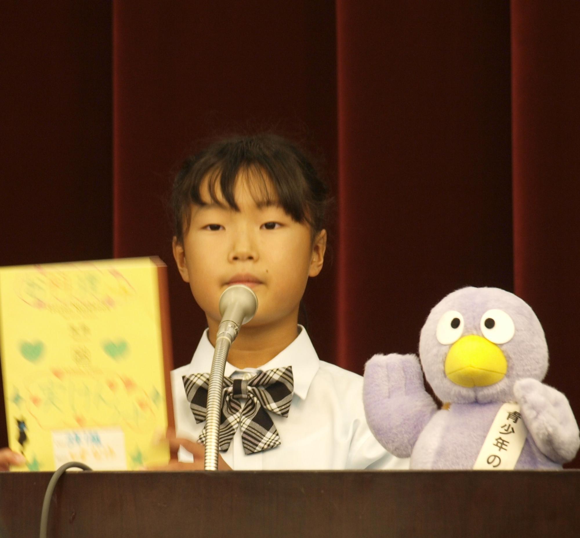 コバトンの置かれた壇上で黄色い本を持ち、発表している様子の新和小 石倉なゆさんの写真