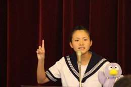 コバトンのマスコット人形がある演台のマイクで発表をしている制服を着た女の子が右手をの人差し指を上に向けて力説している写真