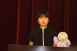 コバトンのマスコット人形がある演台のマイクで発表をしている黒色の服を着た女の子の写真