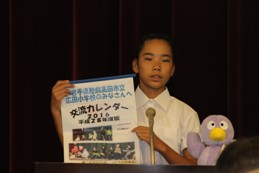 コバトンのマスコット人形がある演台のマイクで交流カレンダーを両手で持ち発表をしている制服を着た男の子の写真
