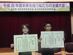 平成26年度 未来を担う私たちの主張大会と書かれた横断幕を背に、2人の児童が両手に賞状を持っている様子の写真