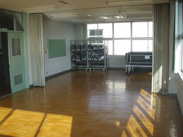 たくさんの折りたたみ椅子が収納された可動式のラックと、折りたたんだ長机が窓際に寄せられている、アコーディオンカーテンの仕切りが付いたフローリング床の講習室の写真