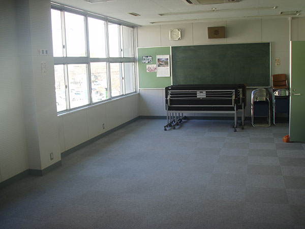 壁に黒板と掲示板があり、その前に畳んだ長机と折りたたみ椅子が寄せて置かれている、大きな窓のある、白い防音壁とグレーのカーペットの床の音楽室の写真