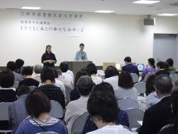上部に「子どもと本との幸せな出会い」と書かれた部屋で、正面に立って話をしている2人の関係者と、椅子に座っている参加者達の写真