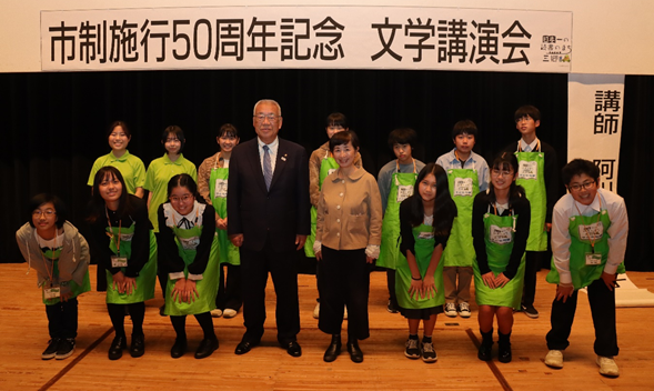 吊り看板に「市制施行50周年記念 文学講演会」と書かれた舞台上で、木津市長と阿川佐和子氏を中心に緑のエプロンを着た子ども司書たちが並んで記念撮影をしている写真