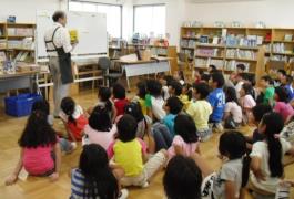 図書館内の一室で、たくさんの子ども達が座って見ている中、エプロン姿の関係者一人が正面に立って話をしている写真